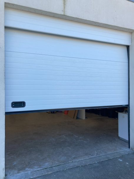 Porte de garage blanche partiellement ouverte, avec mur adjacent en béton.