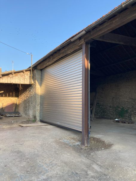 Porte de garage enroulable ouverte dans un bâtiment rural. Mur en pierre à côté.