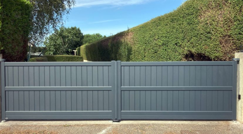 Portail en métal gris, fermé, entouré de haies verdoyantes sous un ciel bleu.