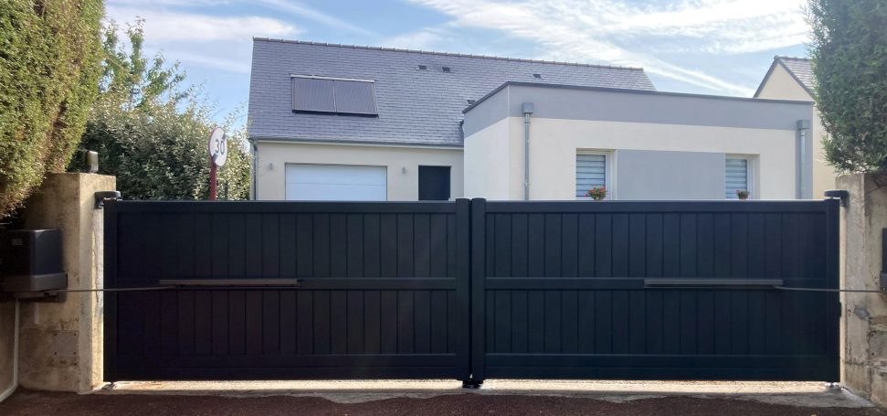 Maison moderne avec un portail noir, entourée de verdure et sous un ciel clair.