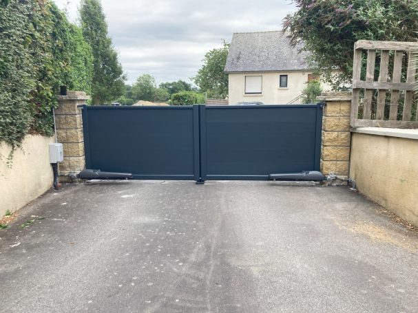 Portail noir fermé avec des murs en pierre et des arbres alentours.