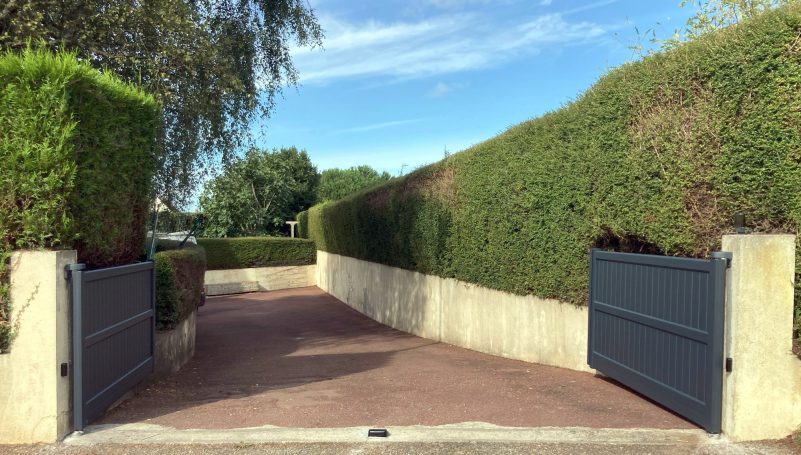 Entrée d'une allée bordée de haies, avec portails gris ouverts sous un ciel bleu.